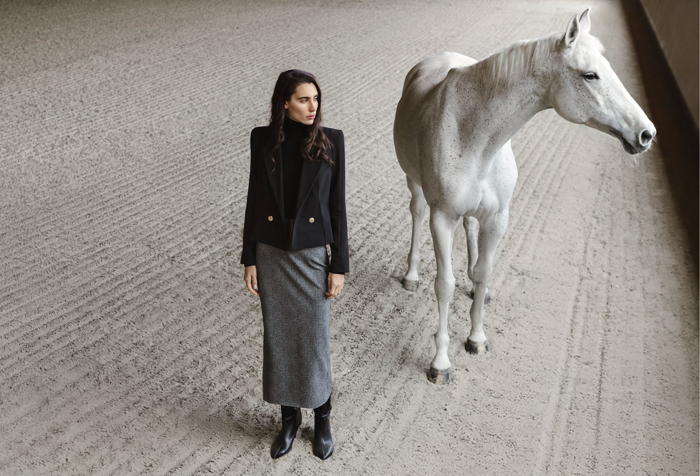 Woman in the TROINA black short blazer and the TORRONA checkered midi-skirt standing next to a white horse.