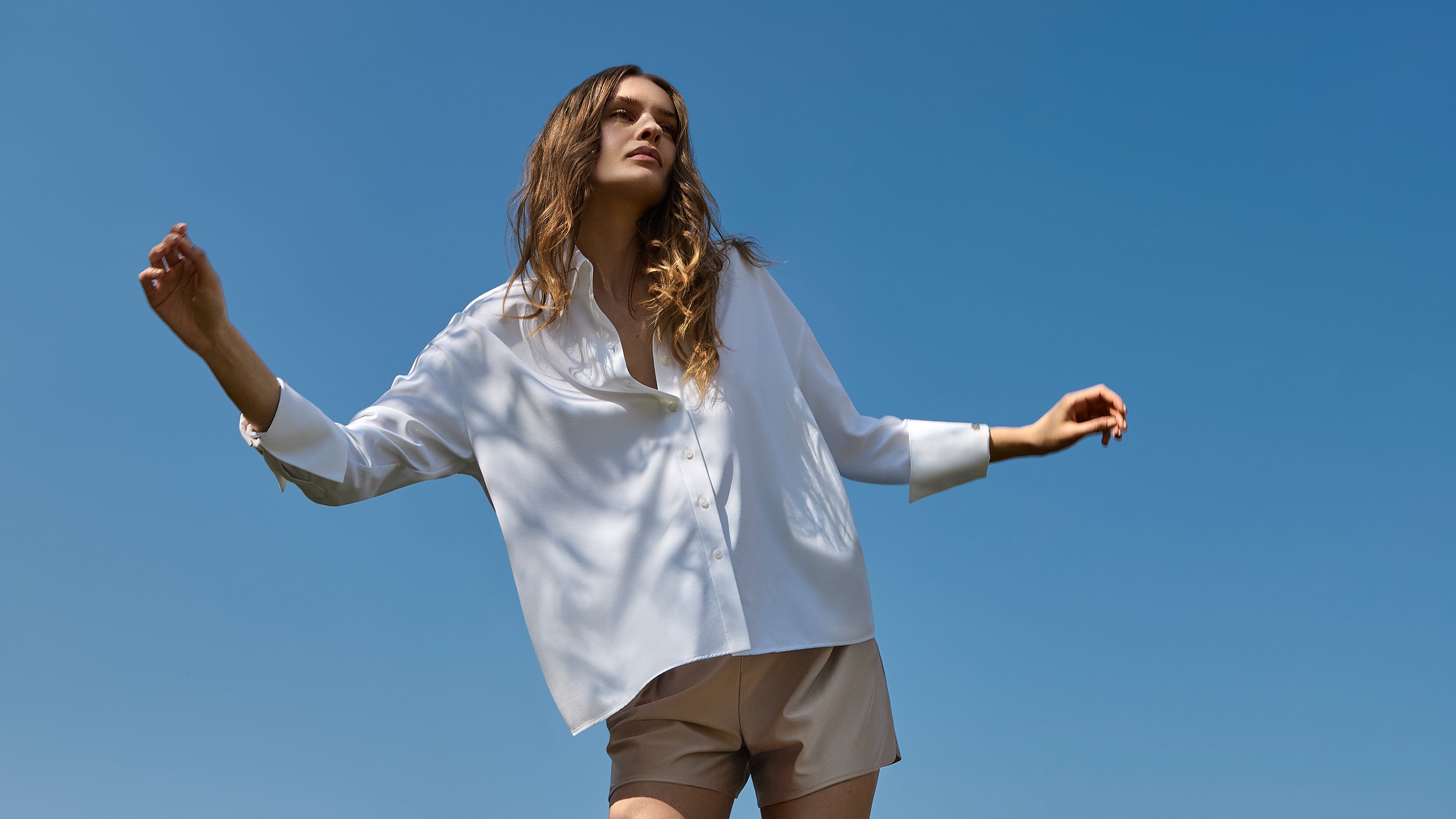 Model in the white INA KESS LAREGIA shirt and golden coloured shorts standing against a clear blue sky