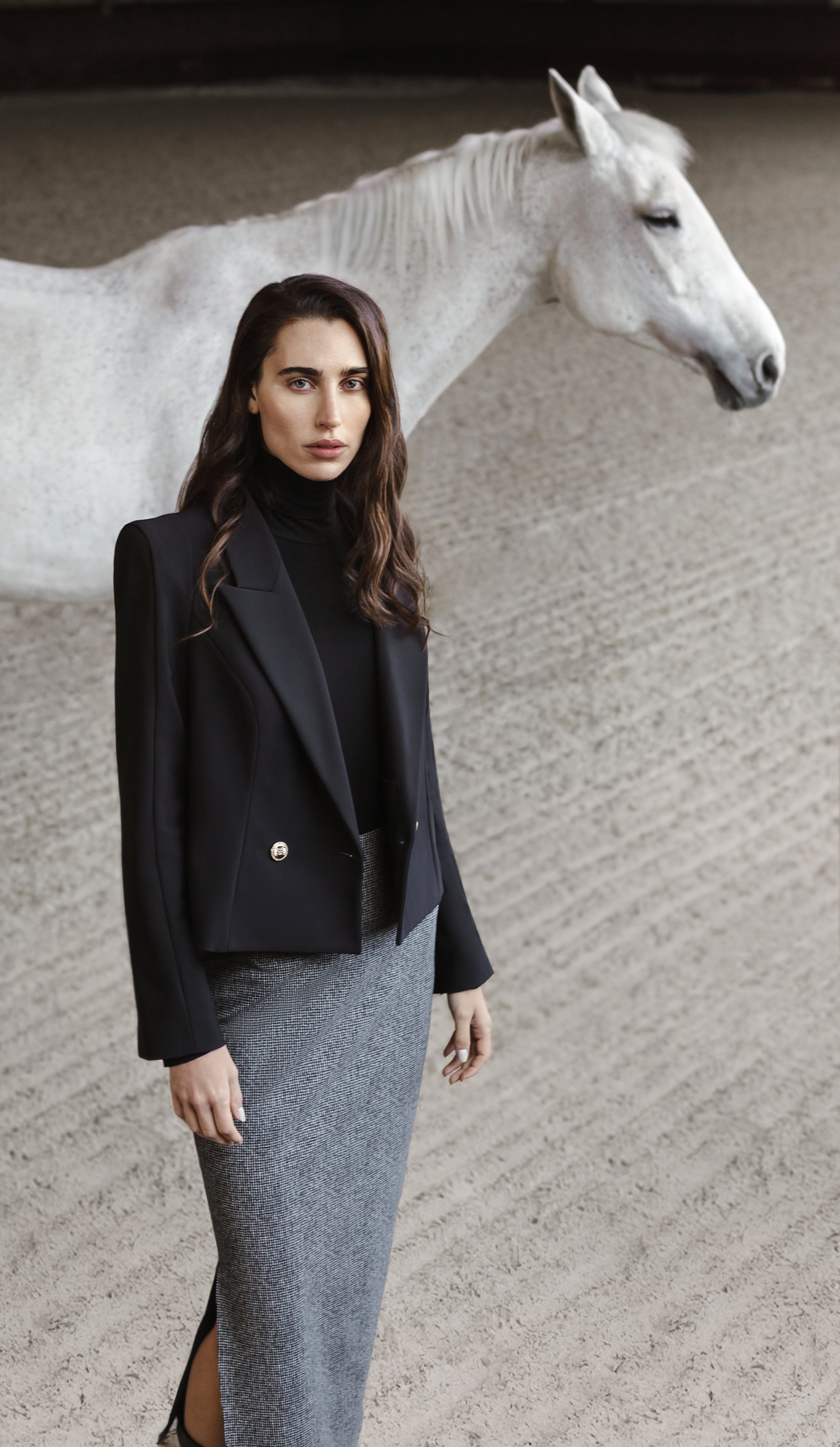 Woman in the TROINA black short blazer and the TORRONA checkered midi-skirt standing next to a white horse.