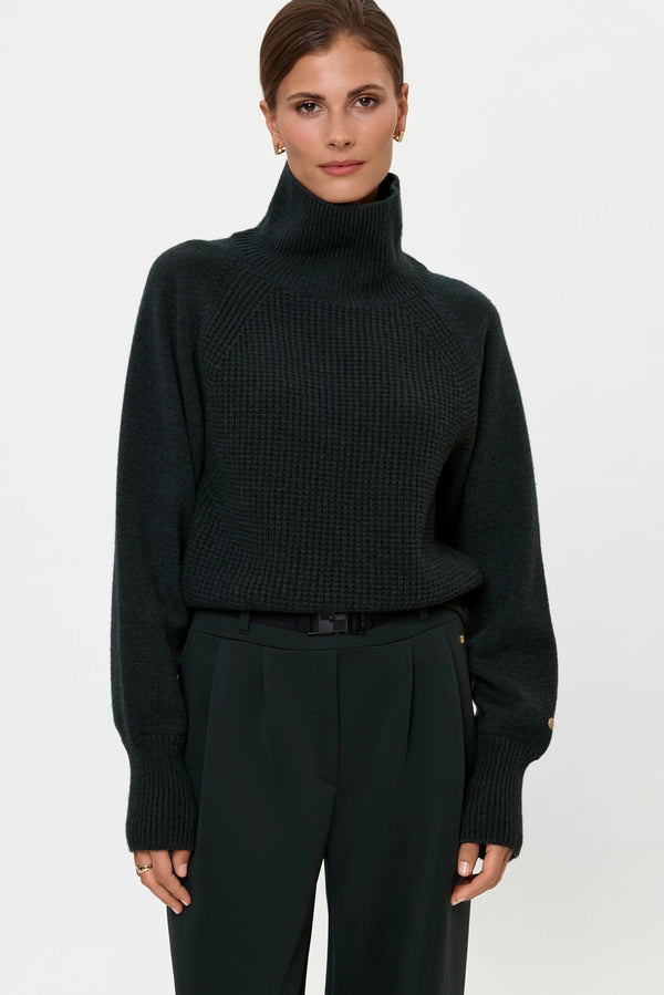 Model wearing a forest green cashmere turtleneck tucked into pleated forest green stretch pants with a black belt