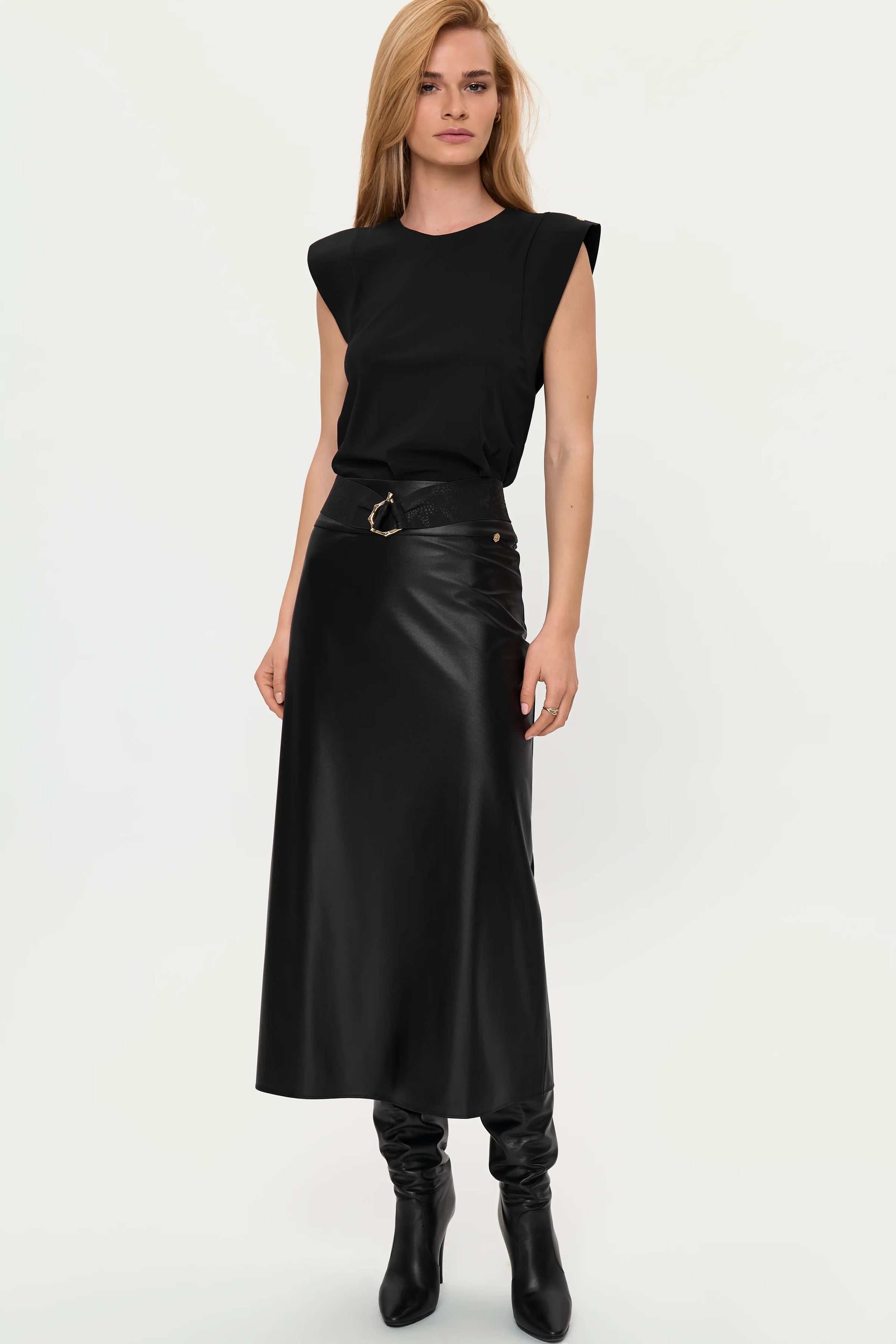 Full-length front view of the VITA Faux Leather Skirt in Black styled with the ERI Top in Black and LEO Belt, highlighting the glossy midi skirt, belted waist, structured shoulder top, and black heeled boots.