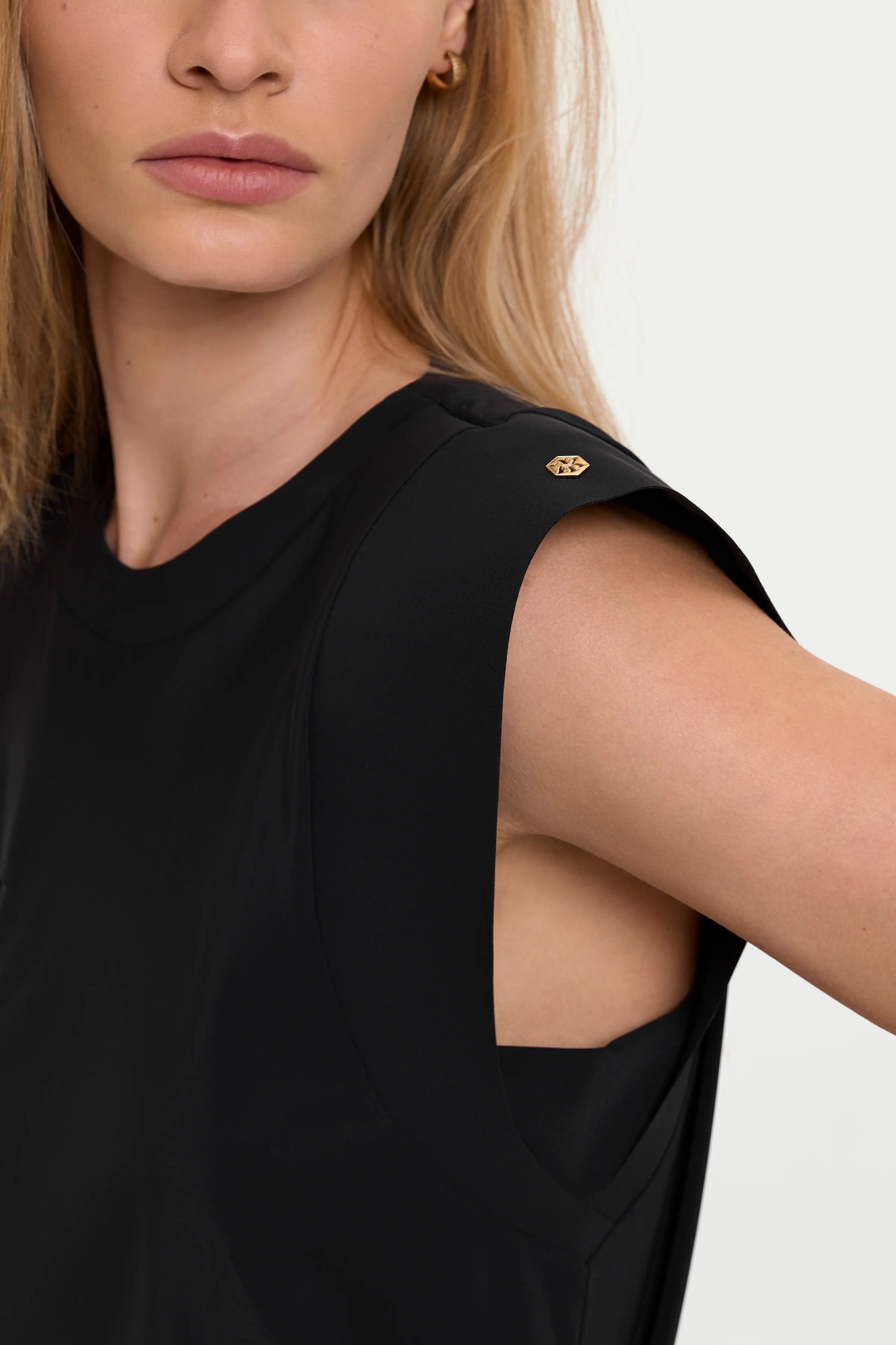 Close-up detail of the ERI Top in Black, showing the sleeveless padded-shoulder design, round neckline, soft drape, deep armhole, and small gold logo at the shoulder.